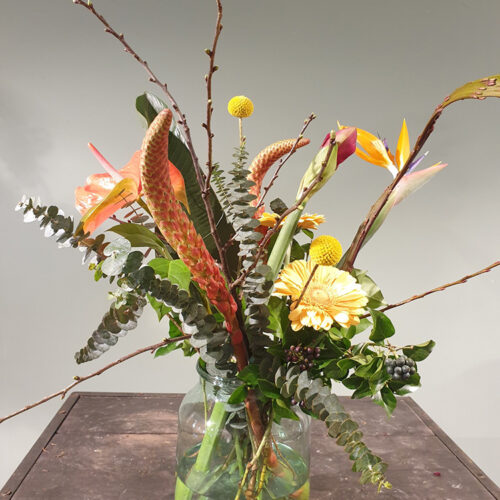 Een stijlvol boeket in een glazen pot met oranje en gele bloemen en verschillende groene takken